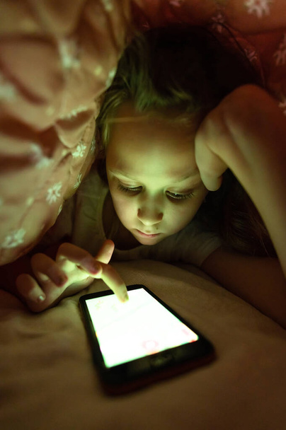 Little girl at night with a smartphone under the covers. Childhood gadget addiction and insomnia - 写真・画像