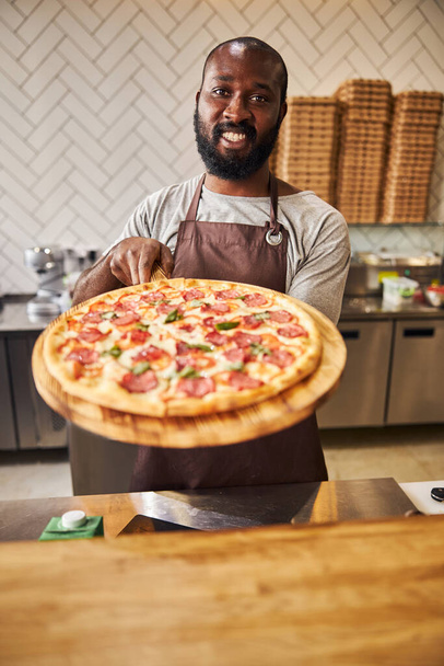 Alegre hombre afroamericano sosteniendo tablero de madera con pizza - Foto, Imagen