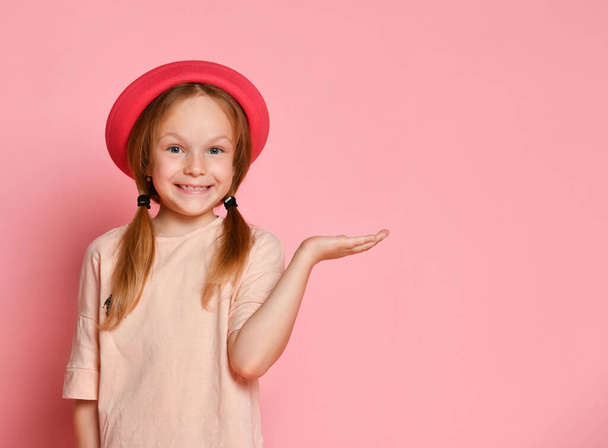 Criança loira de chapéu e t-shirt. Ela está sorrindo e agindo como segurando algo na palma da mão. Posando no fundo do estúdio rosa. Fechar - Foto, Imagem