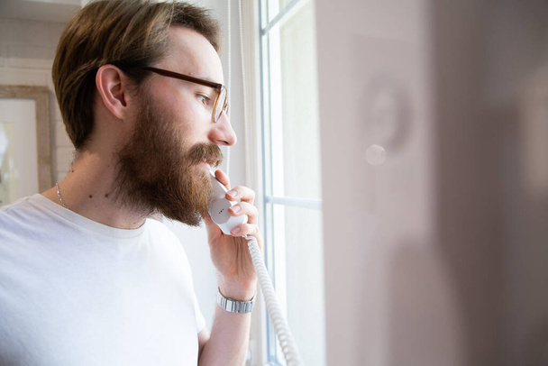 A young bearded man with glasses in a white t-shirt holding a household intercom receiver in a bright interior and looking out the window. Picking up a receiver to open the door with an entryphone. - Φωτογραφία, εικόνα