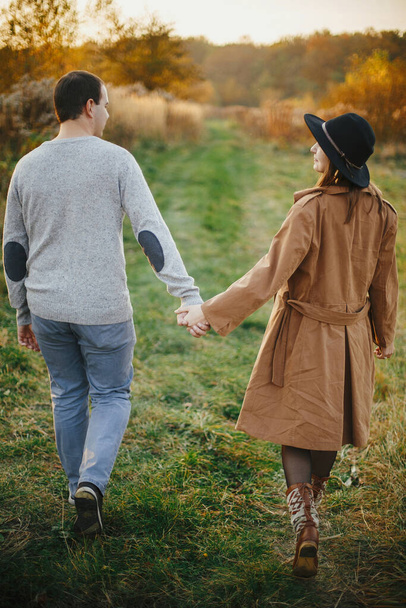 Ευτυχισμένο κομψό ζευγάρι περπάτημα κρατώντας τα χέρια το φθινόπωρο λιβάδι σε ζεστό φως ηλιοβασίλεμα. Ρομαντική αισθησιακή στιγμή. Νεαρή μοντέρνα γυναίκα και ο άνθρωπος χαλάρωση στον τομέα του φθινοπώρου - Φωτογραφία, εικόνα