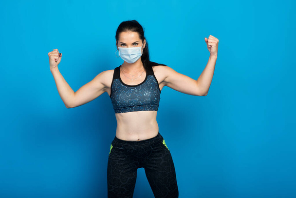 Νεαρή δυνατή γυμνάστρια με μάσκα προσώπου που γυμνάζεται απομονωμένη σε στούντιο με φόντο τον μπλε τοίχο. - Φωτογραφία, εικόνα