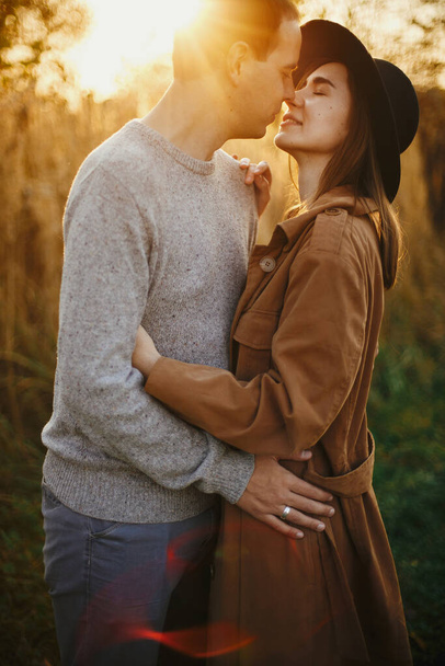 Ευτυχισμένο κομψό ζευγάρι αγκαλιάζει το φθινόπωρο λιβάδι σε ζεστό φως ηλιοβασίλεμα. Ρομαντική αισθησιακή στιγμή. Νεαρή μοντέρνα γυναίκα και ο άνθρωπος φιλιά στον τομέα του φθινοπώρου - Φωτογραφία, εικόνα