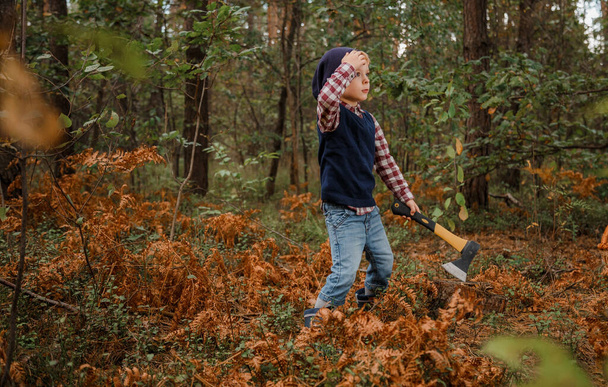 Kisfiú az őszi erdőben. Egy fiú baltával a fa mellett. A favágó tartja a fejszét. Fejszével áll az erdőben.. - Fotó, kép