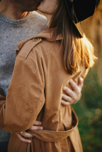 Sonbahar çayırlarında sıcak gün batımı ışığında kucaklaşan şık bir çift. Nazikçe sarılıyorlar. Romantik duygusal an. Genç adam kahverengi ceketli, şık bir kadına sarılıyor. - Fotoğraf, Görsel
