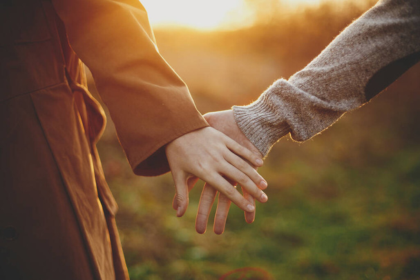 Az érzéki pár gyengéden fogja egymás kezét az őszi rét meleg napnyugtakor. Közelkép a fiatal férfiról és nőről, amint napsütésben fogják egymás kezét. Romantikus együttlét pillanat, konceptuális kép - Fotó, kép