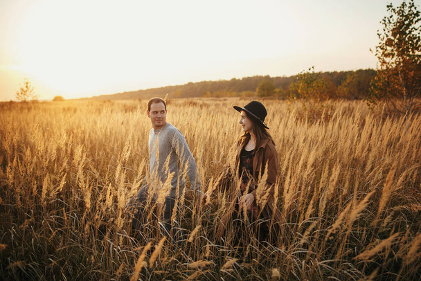 Stílusos pár sétál a meleg naplemente fény őszi területen. Fiatal, divatos nő és férfi kéz a kézben a fű és gyógynövények között a napsütésben. Romantikus autentikus pillanat, utazás együtt - Fotó, kép