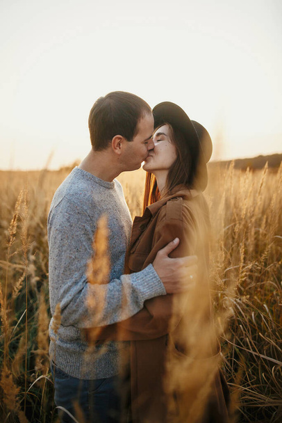 秋のフィールドで暖かい夕日の光を受け入れるスタイリッシュなカップル。若いファッショナブルな女性と男は太陽の下で草やハーブの間で抱き合ってキスをします。ロマンチックな本物の瞬間 - 写真・画像