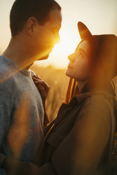 秋のフィールドで暖かい夕日の光に包まれたスタイリッシュな官能的なカップル。若いファッショナブルな女性と男性が優しく太陽の光を背景に抱擁。ロマンチックな本物の瞬間 - 写真・画像