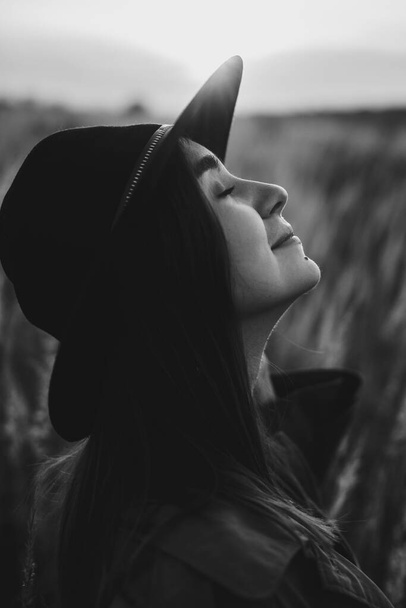 Portrait of calm boho woman in  sunset light in autumn field. Young stylish female in hat posing on background of grass and herbs in sunshine. Sensual moment, black and white photo - Fotografie, Obrázek