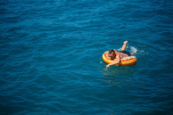 Nuori mies kelluu puhallettavalla ilmarenkaalla meressä sinisellä vedellä. Juhlava loma onnellisena aurinkoisena päivänä. - Valokuva, kuva