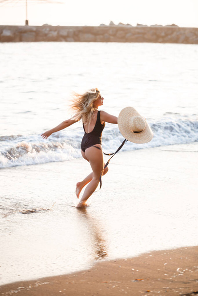 Bella ragazza in costume da bagno nero e cappello su una spiaggia di sabbia al mare alla luce del sole del tramonto - Foto, immagini