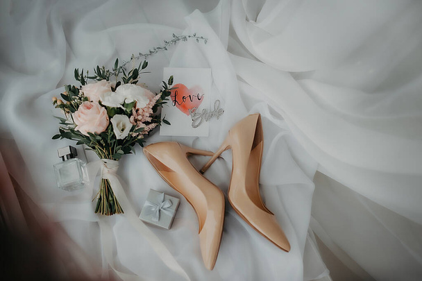 stílusos kiegészítők a menyasszony öltözködési és esküvői ruha, bézs lakk bőr cipő, ezüst doboz gyűrűk és egy elegáns csokor bazsarózsa rózsa fehér tüll háttér - Fotó, kép
