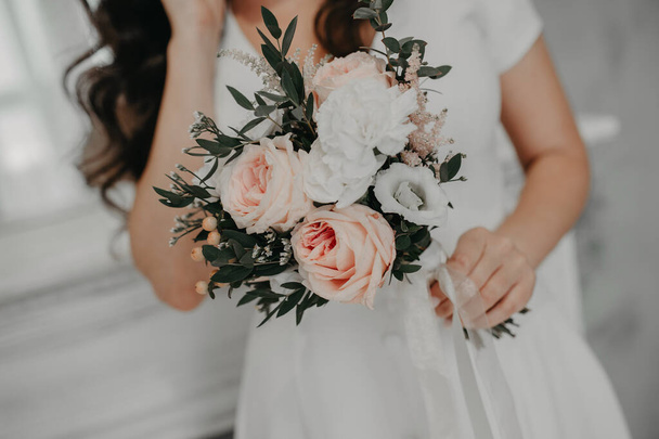 стильная невеста без лица в скромном простом белом платье держит невероятный букет розовых пыльных цветов из пионских роз и зеленых растений для украшения - Фото, изображение