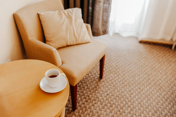 elegante interior en colores cálidos, una silla fácil con una almohada y utensilios de té blanco en la mesa, un lugar para una oficina en casa o un lugar para relajarse - Foto, Imagen