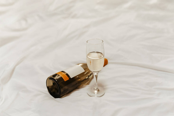 a bottle of wine and a beautiful glass goblet on the bed with pure white crumpled pastel linen in the dim light of lamps on a date or a romantic evening - Photo, Image