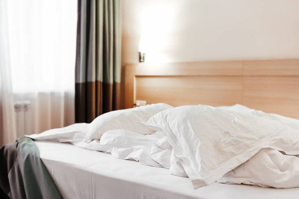 мятая белая фабрика постельных принадлежностей на кровати с одеялом и подушками, рядом с прикроватным столиком и темными тяжелыми шторами на полу, стандартный элегантный вид гостиничного номера - Фото, изображение