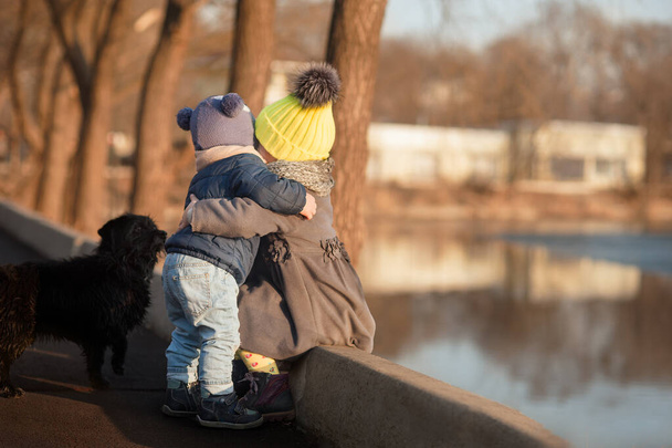 een jongen en een meisje warm gekleed in jassen en gekleurde hoeden met een zwarte hond in de buurt stevig knuffelend tegen de achtergrond van het water, een kaart voor Valentijnsdag over familie en relaties - Foto, afbeelding
