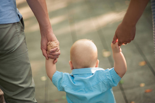 Родители учат ребенка ходить, крепко держат его за руку с обеих сторон, ребенок в голубой рубашке и без волос, вид сзади - Фото, изображение