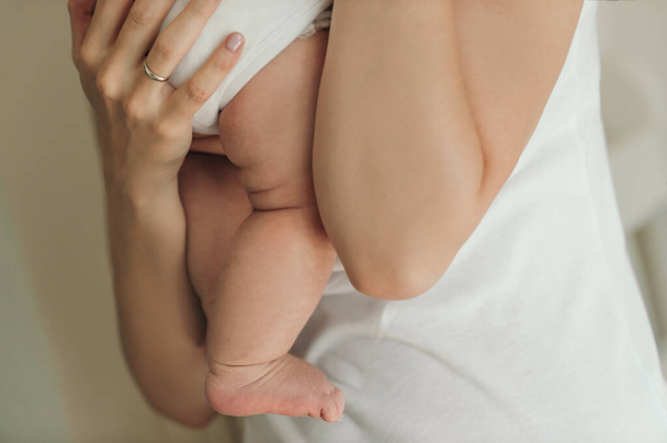 μητέρα με λευκά ρούχα αγκαλιάζει το μικρό νεογέννητο μωρό της με γυμνά παχουλά πόδια με πτυχώσεις πολύ σφιχτά και ευλαβικά σε ένα απλό μπεζ φόντο - Φωτογραφία, εικόνα