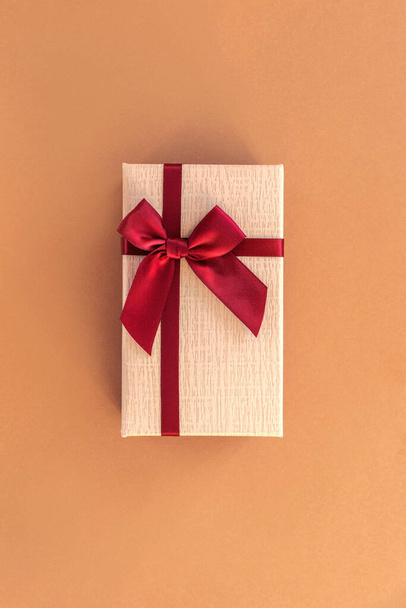 Cadeau dans une boîte avec noeud rouge foncé sur fond beige, vue de dessus. Pose plate - Photo, image
