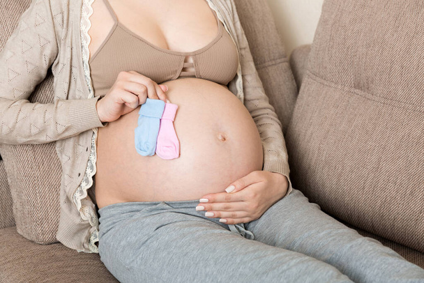beautiful pregnant young woman holding a pair of cute baby socks on her tummy, lying on sofa at home. - Zdjęcie, obraz