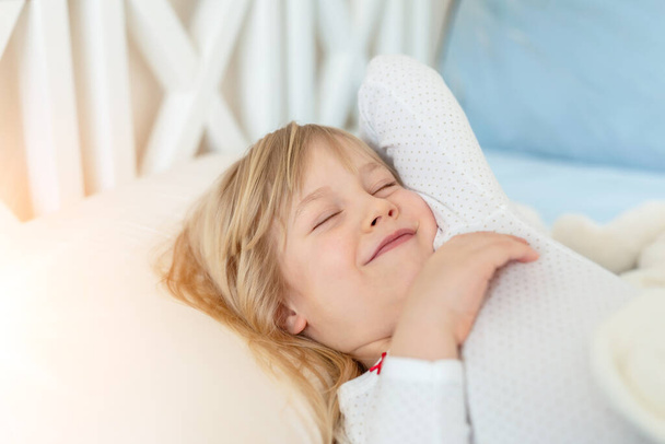 Cute adorable caucasian blond little toddler kid girl lying in bed on white pillow. Adorable sweet happy smiling child stretching wake up early morning for kindergarten or school. Childhood concept. - 写真・画像