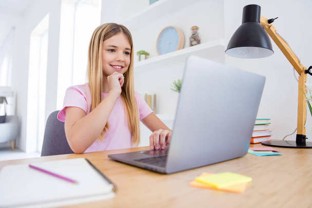 Photo of positive kid girl sit table desk study remote use laptop read watch education video webinar touch hand chin in house indoors - Valokuva, kuva