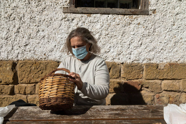 A senior adult woman, wearing a face mask, searches for something in a wicker basket before eating in front of the mountain hut of the fountain of Artica, in Luesia, Spain. - Photo, Image
