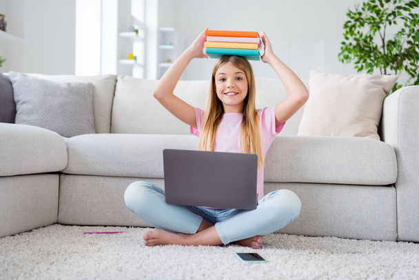 Full length photo of positive kid girl study remote laptop hold pile stack books lesson materials head sit floor legs crossed folded in comfort cozy house indoors - 写真・画像