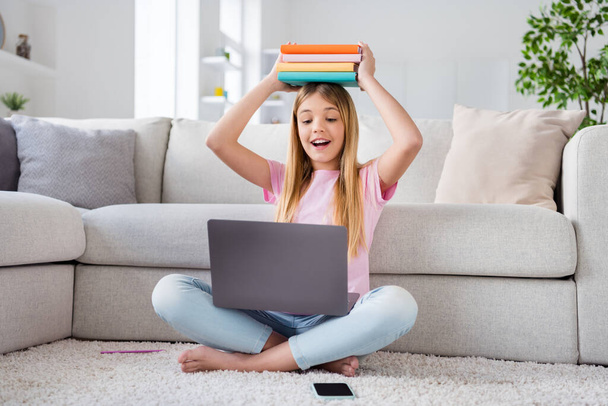 Full length photo of excited small kid girl work remote laptop have online tutor lesson meeting hold pile stack books head sit floor legs crossed folded in house indoors - Foto, Imagen