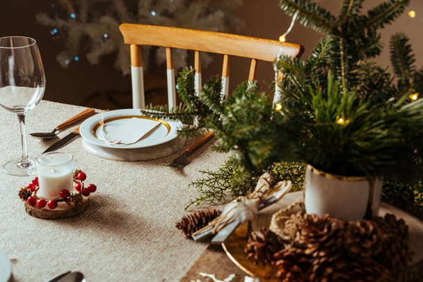 Homely christmas table setting, decorated with pine branches , pine cones, and rustic tablecloth surrounded by Nordic decoration and lights, with the illuminated tree in the background. Family dinner. - Фото, изображение