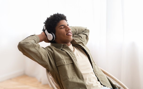 alegre negro adolescente chico en auriculares relajante a su favorito música mientras sentado en cómodo sillón, panorama - Foto, Imagen
