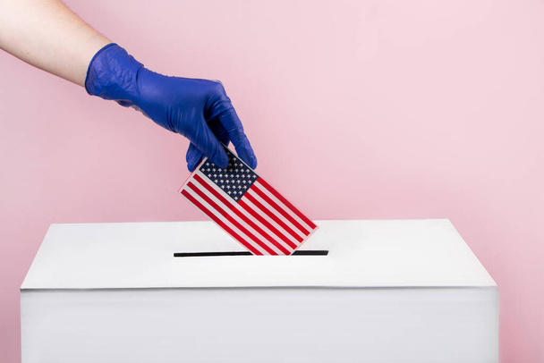 Женщина в синих защитных перчатках кладет флаг США в урну для голосования. Концепция выборов и коронавируса. - Фото, изображение
