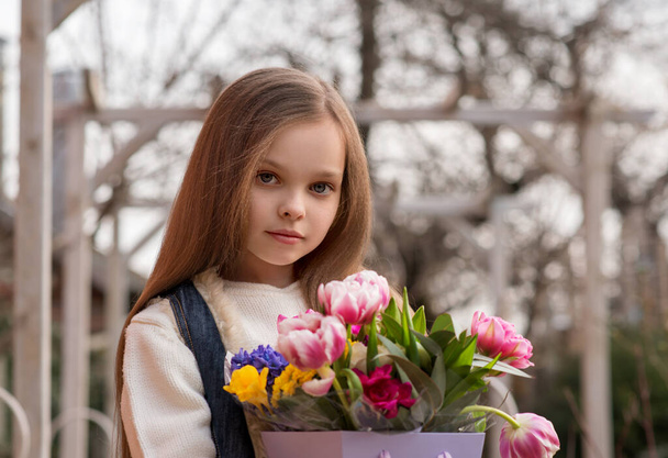 Cute smiling little girl with bouquet of spring flowers in hands outdoor. Portrait of kid with long hair with on rural background with seasonal floral gift. Concept of Easter or Mother Day celebration. - Photo, image