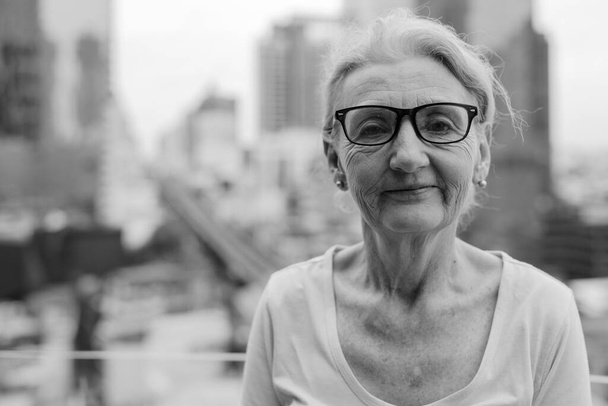 Портрет красивой пожилой женщины с светлыми волосами с видом на город в черно-белом цвете - Фото, изображение