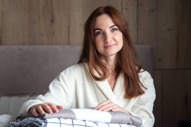 Young woman with beautiful smile in warm beige robe is leaning on stack of folded bed sheets in different texture. Bedding from natural and organic cotton. Copy space. Manufacture. Hotel industry. - Fotografie, Obrázek