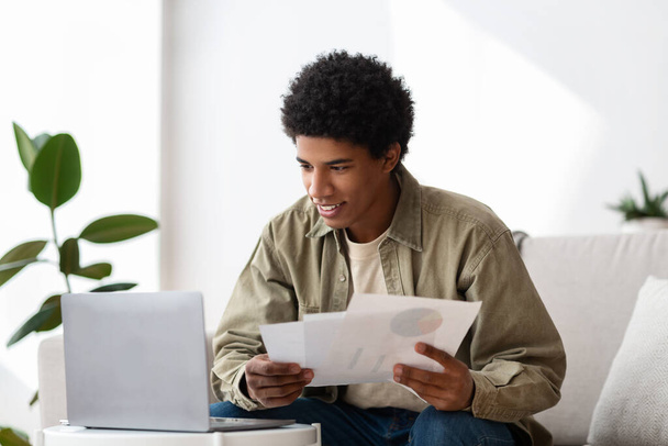 Chico negro positivo con portátil que estudia en línea en casa, escribiendo papel de curso o preparándose para el examen - Foto, Imagen