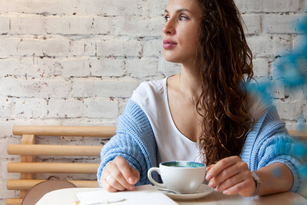 Ευτυχισμένη νεαρή γυναίκα σε πλεκτό ζεστό πουλόβερ κρατώντας με τα χέρια ένα φλιτζάνι υγιεινό ζεστό μπλε καφέ latte. Γαλάζιος καφές latte φτιαγμένος με φρέσκους κόκκους καφέ και bluechai τσάι μπιζελιού πεταλούδας. Ευεξία έννοια. - Φωτογραφία, εικόνα