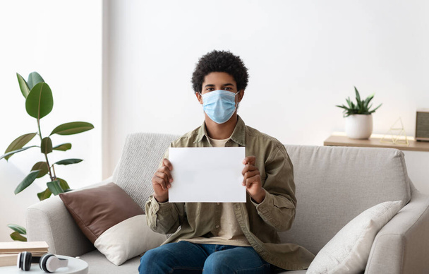Estudante masculino preto em máscara médica segurando sinal de papel em branco dentro de casa, espaço para o seu projeto - Foto, Imagem