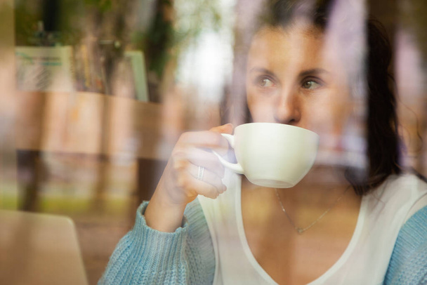View through window of young woman wearing blue sweater talking on phone while working on computer and enjoying cup of hot drink in coffee shop. Street reflection. Freelance concept. - Photo, image