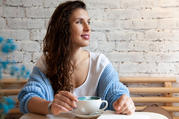 Happy young woman in knitted warm sweater holding with hands a cup of healthy hot blue coffee latte. Blue coffee latte made with fresh coffee beans and bluechai butterfly pea tea. Wellbeing concept. - Fotografie, Obrázek