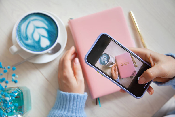 Woman taking photo of blue coffee latte, pink coloured planner 2021 and gold pen on her smartphone. People and technology. Taking picture for posting and sharing on social media. Blogging concept. - Foto, imagen