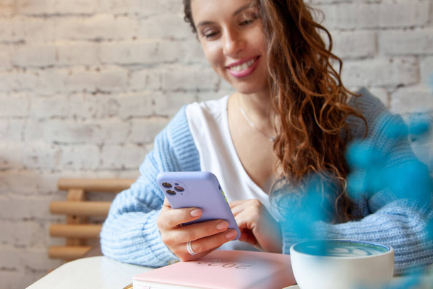 Egy fiatal nő kék meleg pulóverben SMS-t olvas a mobiljáról, miközben kávét iszik. Üzletasszony ellenőrzi a közösségi médiát az okostelefonján. Blogger életmód. Tervezés és szervezés - Fotó, kép