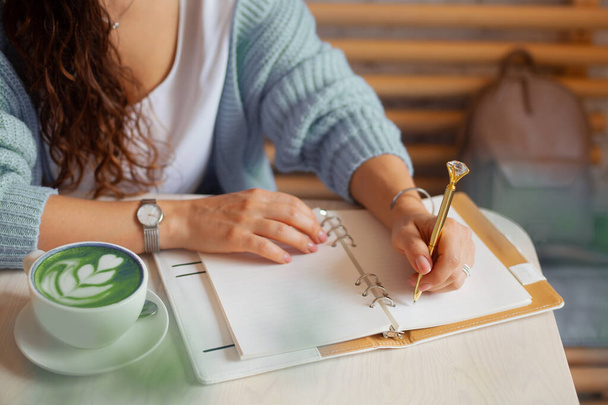 Well groomed woman hand holding gold pen and writing notes with gold pen in notebook while drinking blue latte beside window. Freelance journalist working at home. Planning future concept. Copy space - Photo, image