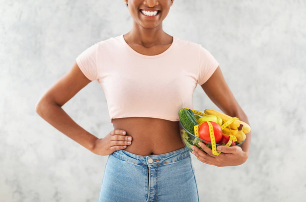 Vista de cerca de una joven afroamericana sosteniendo frutas y verduras frescas con cinta métrica sobre fondo gris - Foto, imagen
