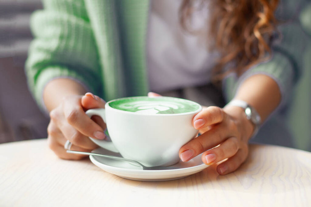 Unfocused woman hands holding cup of hot butterfly pea latte or blue spirulina latte on wooden table. Organic healthy and trendy drink. Wellbeing and detoxing concept. Copy space. - Фото, изображение