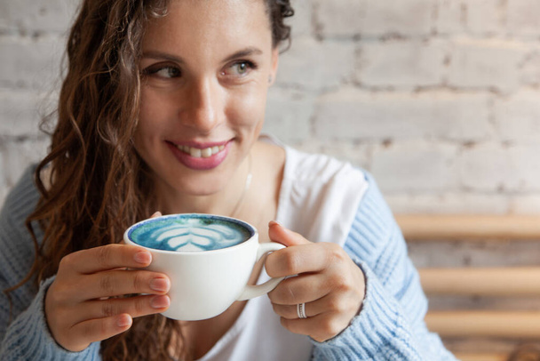 Happy young woman in knitted warm sweater holding with hands a cup of healthy hot blue coffee latte. Blue coffee latte made with fresh coffee beans and bluechai butterfly pea tea. Wellbeing concept. - Фото, изображение