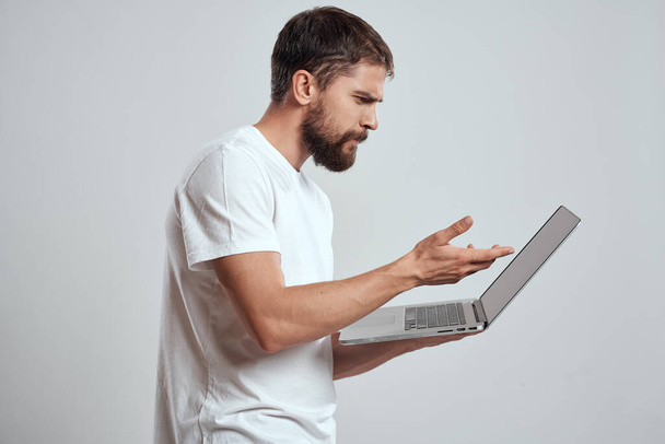 Ένας άνθρωπος με ένα φορητό υπολογιστή στα χέρια του σε ένα ελαφρύ φόντο σε ένα λευκό t-shirt συναισθήματα φως φόντο νέες τεχνολογίες - Φωτογραφία, εικόνα