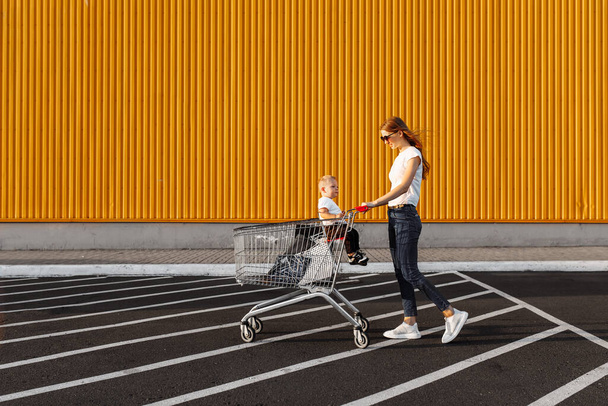 Attractive happy young woman in sunglasses shopping with small child, trolley and shopping bags, outdoors - Zdjęcie, obraz